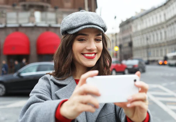 Mujer encantadora en elegante sombrero gris y abrigo haciendo selfie mientras camina más allá del viejo edificio. —  Fotos de Stock