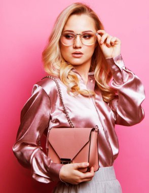 Bluz ve pantolon giyen, gözlüklü, pembe arka planda duran güzel sarışın kadın..