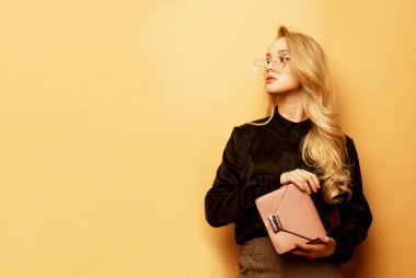 Kahverengi bluzlu ve pantolonlu genç sarışın kadın pembe bir el çantası tutuyor ve bej bir arka planda poz veriyor. Moda ve insanlar konsepti..