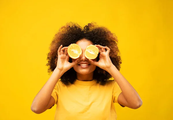 lifestyle, food, diet and people concept: portrait of young afro woman holding a pair of half oranges covering her eyes. African woman holding fresh fruit on yellow background.