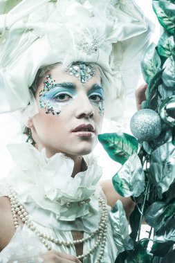 Parti, Noel ve insanlar: Noel karnavalı için kostüm giymiş güzel bir kadın Noel süslemeleri ve gümüş yapraklar çerçevesinde poz veriyor. Yaratıcı makyaj, saç stili.