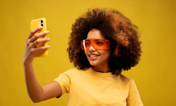 lifestyle, tehnology and people concept: Close up young woman of African American ethnicity wears yellow t-shirt do selfie shot pov on mobile phone isolated on yellow background.