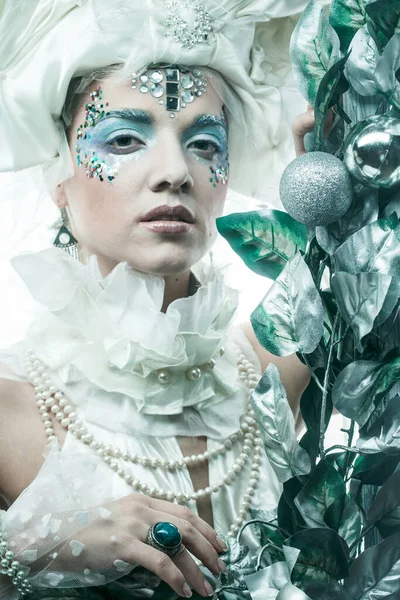 Parti, Noel ve insanlar: Noel karnavalı için kostüm giymiş güzel bir kadın Noel süslemeleri ve gümüş yapraklar çerçevesinde poz veriyor. Yaratıcı makyaj, saç stili.