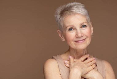 Elleri yüzlerinde gülümseyen olgun bir kadının güzel portresi. Yaşlanma karşıtı tedaviden sonra kendini zinde hisseden mutlu son sınıf kadınının yakın çekimi. Gülümseyen güzel kusursuz ciltli kameraya bakıyor..