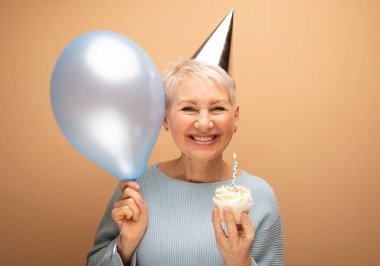 İnsanlar, parti, yaşlanma ve olgunluk kavramı. Doğum gününü kutlayan koni şapkalı, elinde mum ve mavi balonla yalnız poz veren güzel, yaşlı bir kadın. Mutlu zamanlar..