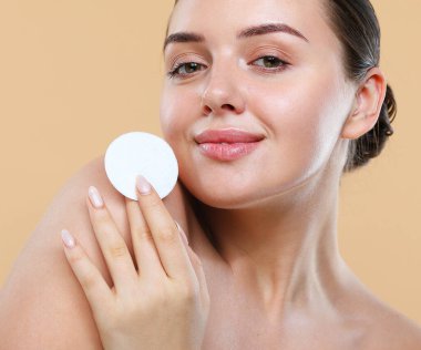 Makyajı kaldıran genç bir kadın, yüzüne pamuk pedleri tutuyor. Bej arka planda mükemmel ciltli bir kadının fotoğrafı. Güzellik ve cilt bakımı.