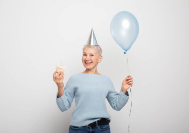 Mutlu yaşlı kadın koni şapkası takıyor doğum gününü mum ve gri arka planda mavi balonla kutluyor.