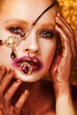 Ağzında değerli bir yüzük tutan genç ve güzel bir kadın. Kolye gözlerini kapatıyor. Parlak makyaj, dudaklarında altın. Altın arka planda poz veren model kadın..
