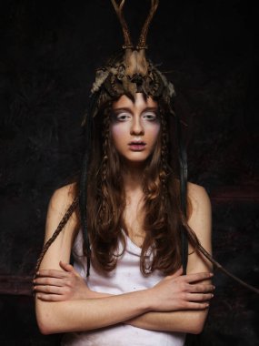 Parlak makyajlı, örgülü ve kafasında kafatası olan genç bir kadın kollarını siyah bir arka planda kavuşturmuş duruyor.