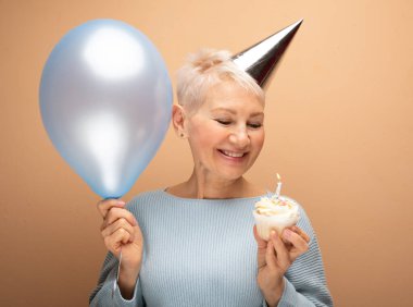 İnsanlar, parti, yaşlanma ve olgunluk kavramı. Doğum gününü kutlayan koni şapkalı, elinde mum ve mavi balonla yalnız poz veren güzel, yaşlı bir kadın. Mutlu zamanlar..