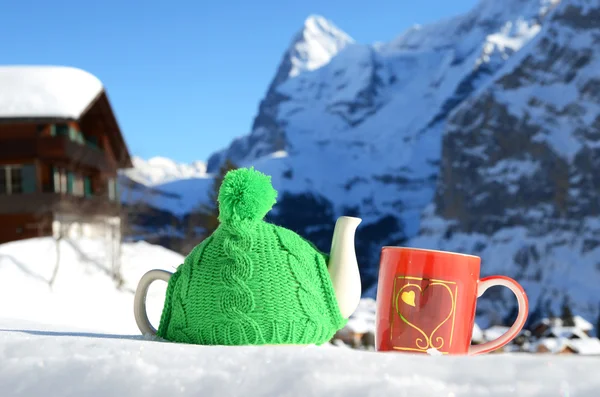 Panela de chá na tampa de malha e xícara vermelha com um coração na neve — Fotografia de Stock