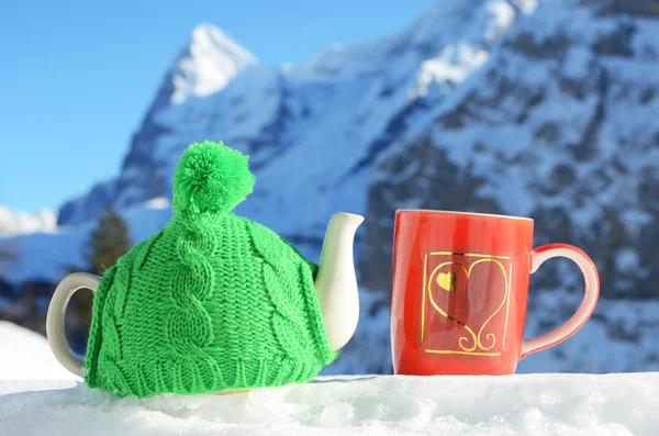 Tekanna i Stickad mössa och röd kopp med hjärtan i snön — Stockfoto