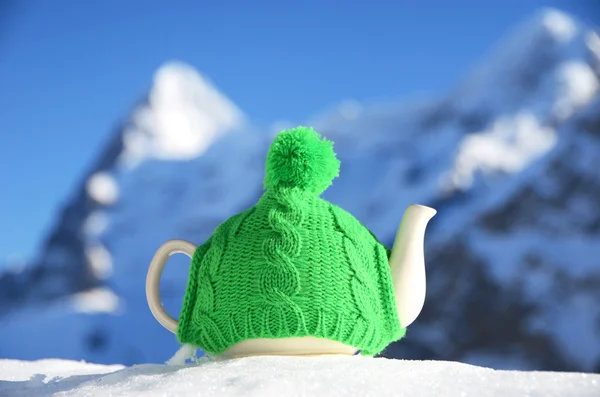 Panela de chá no gorro de malha na neve contra o pico da montanha — Fotografia de Stock