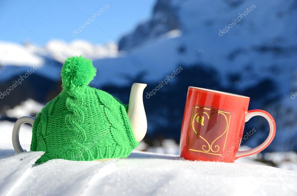 Tea pot in the knitted cap and red cup with a heart in the snow