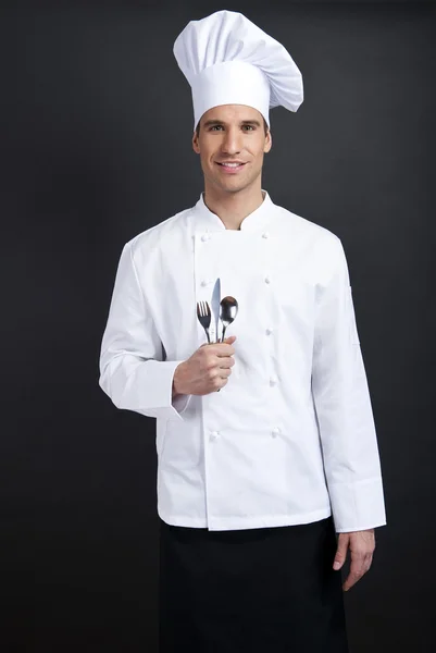 Chef cozinheiro contra fundo escuro sorrindo com chapéu colher holdinf — Fotografia de Stock