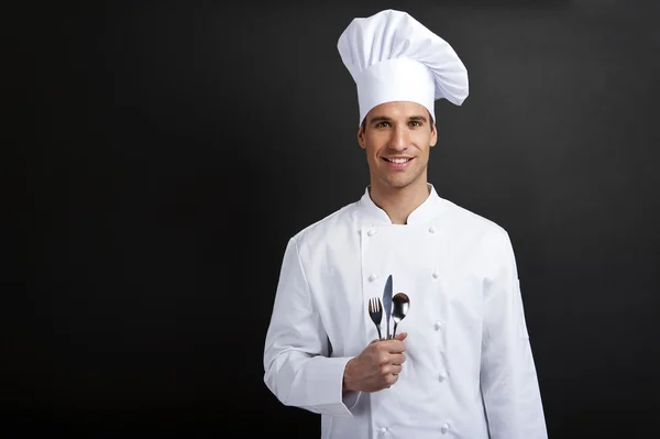 Cuoco cuoco contro sfondo scuro sorridente con cappello holdinf cucchiaio — Foto Stock