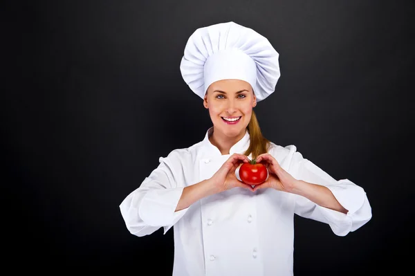 Woman chef make hand heart sign with tomato over dark background — Stock Photo, Image