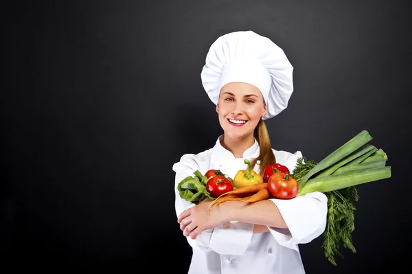 Woman chef holding vegetables and smiling over dark background — Stock Photo, Image