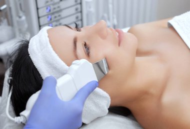 Doktor-kozmetik uzmanı cihazı güzellik salonundaki genç ve güzel bir kadının yüz derisinin ultrasonla temizlenmesi için bir prosedür haline getiriyor. Kozmetoloji ve profesyonel cilt bakımı.