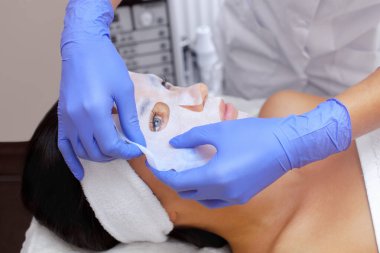 Güzellik salonundaki genç bir kadının yüzüne çarşaf maskesi takmak suretiyle cildi temizleme ve nemlendirme prosedürü. Kozmetoloji ve profesyonel cilt bakımı..