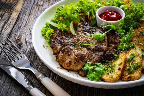 Roasted pork loin and fried potatoes with green vegetables on wooden table