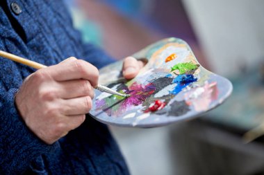 Closeup view of artist painting with palette, brush and acrylic colour paints in art studio indoors