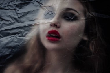 Genç, güzel, üzgün kadın COVID-19 'a karşı korunmak için plastik bir çarşafın arkasında mahsur kaldı. Tam kitap kapağına göre. 