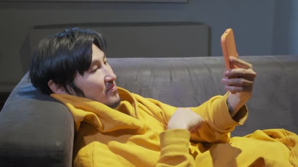 En asiatisk kille lägger sig på soffan på ett litet kontor och kommunicerar via mobiltelefon — Stockvideo