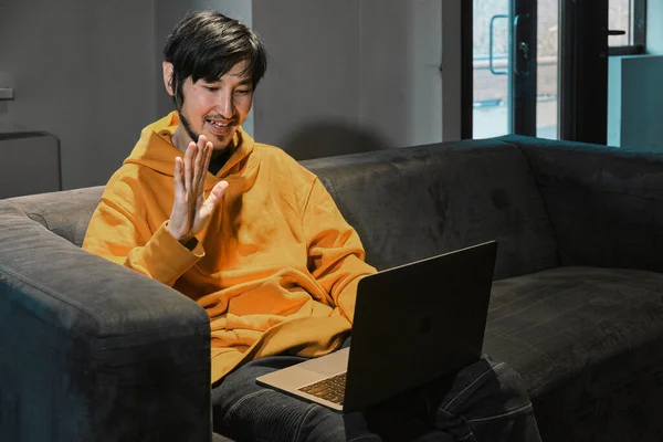 Un mec asiatique est assis sur un canapé dans un petit bureau et communique via un ordinateur portable — Photo