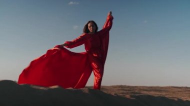Kırmızı elbiseli Asyalı bir kadın kum tepelerinde tek ayak üstünde poz verir.