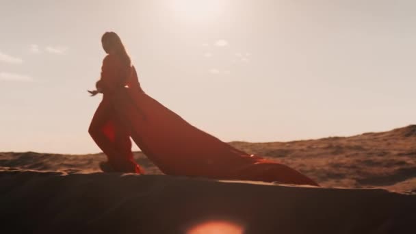 An Asian woman in a red dress dancing against the sun at sunset on sand dunes — Stock Video