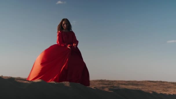 Una mujer asiática en un vestido rojo se pone en una pose en una pierna en dunas de arena — Vídeo de stock