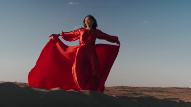 Una mujer asiática en un vestido rojo gira sobre dunas de arena — Vídeos de Stock