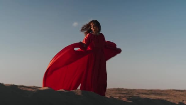 Una mujer asiática en un vestido rojo gira sobre dunas de arena — Vídeos de Stock