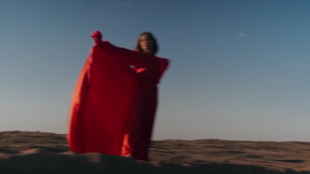 Una mujer asiática en un vestido rojo gira sobre dunas de arena — Vídeos de Stock