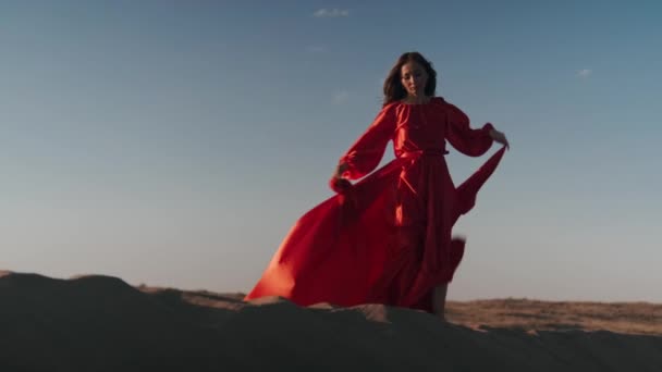An Asian woman in a red dress gets into a pose on one leg on sand dunes — Stock Video