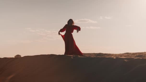 Güneş batarken kum tepelerinde dans eden kırmızı elbiseli Asyalı bir kadın. — Stok video