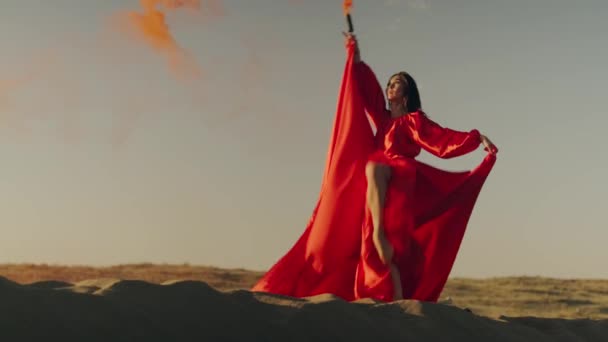Uma mulher asiática em um vestido vermelho com uma bomba de fumaça laranja em suas mãos gira em dunas de areia — Vídeo de Stock