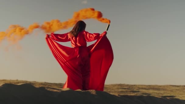 Una asiática vestida de rojo con una bomba de humo naranja en las manos gira sobre dunas de arena — Vídeos de Stock