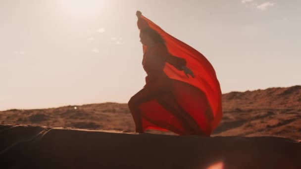 Una mujer asiática en un vestido rojo bailando contra el sol al atardecer en dunas de arena — Vídeos de Stock