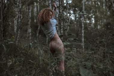 Ormanda çıplak bir kadın, kırmızı kıvırcık saçlı, beyaz bluzla kaplı.