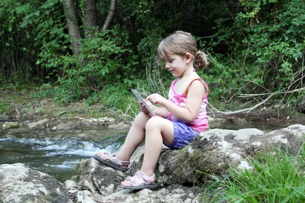 Little girl sitting next to a stream and play with tablet — Stock Photo, Image