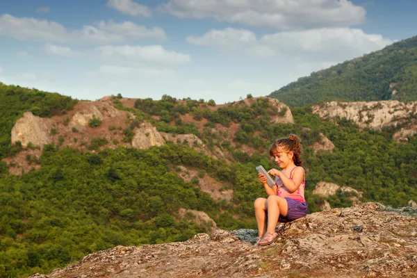 Little girl sitting on a mountain top and play with tablet — Stock Photo, Image