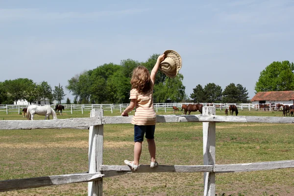 Little girl with hat in hand standing on a corral fence — Stock Photo, Image