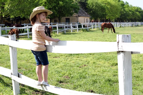 Little girl with cowboy hat standing on corral and watching hors — Stock Photo, Image