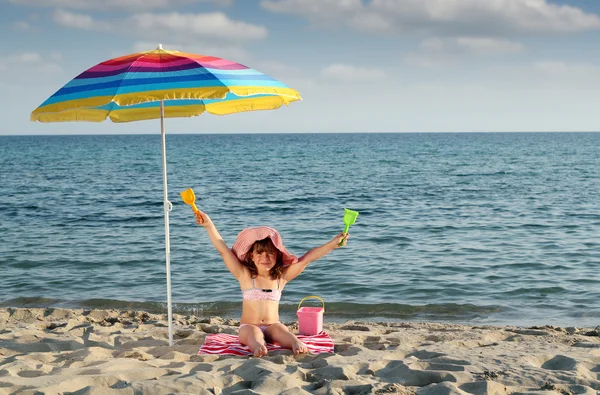 Happy little girl with hands up sitting under sunshade on beach — Stock Photo, Image