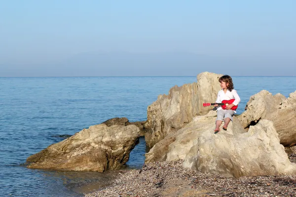 坐在海边岩石上，弹吉他的小女孩 — 图库照片