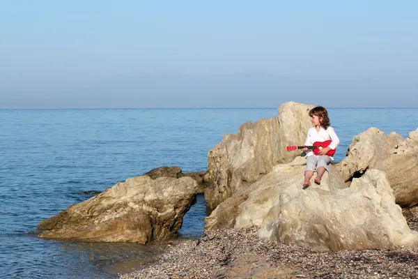 Little girl sitting on a rock by the sea and playing guitar — Stock Photo, Image