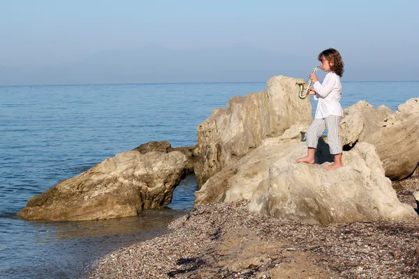 Little girl standing on a rock by the sea and playing saxophone — Stock Photo, Image