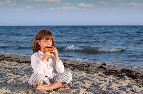 Vacker liten flicka sitter på stranden och spela pan pipe — Stockfoto